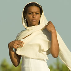 Femme en robe blanche, un foulard crème drapé sur la tête, debout face à un ciel bleu pâle