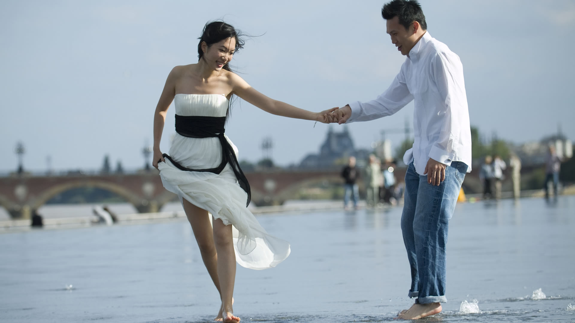 Couple se tenant par la main et marchant pieds nus dans l'eau peu profonde du Miroir d'eau de Bordeaux, elle en robe bustier blanche, lui en chemise blanche et jean