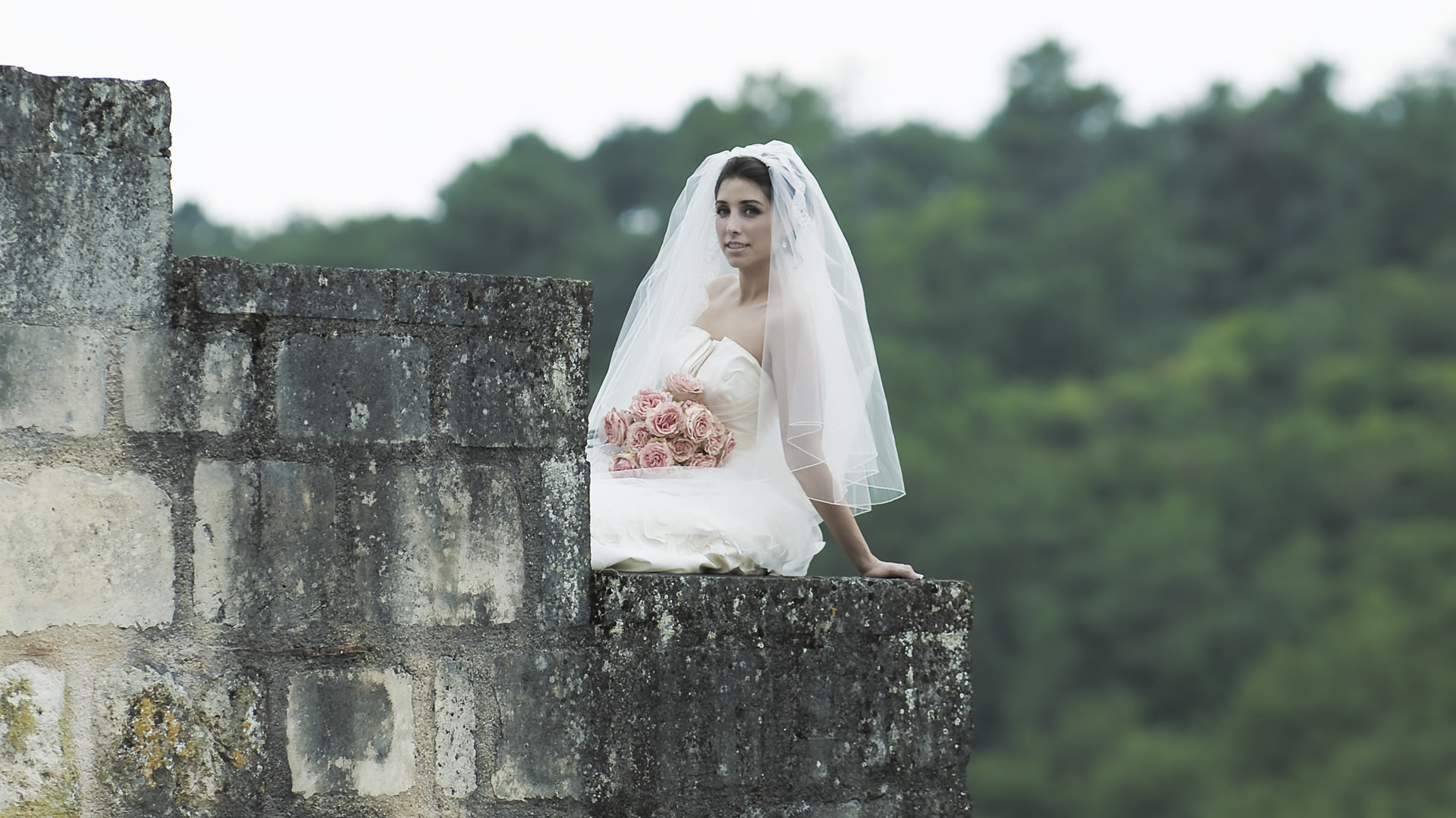 Portrait de mariage artistique d'une mariée en robe blanche et voile, tenant un bouquet de roses roses près d'un vieux mur de pierre