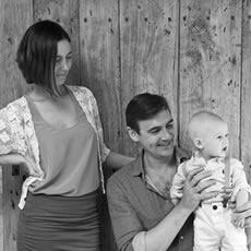 Portrait de famille en noir et blanc de parents avec leur bébé devant un mur en bois rustique – photographie de famille dans le Médoc, France