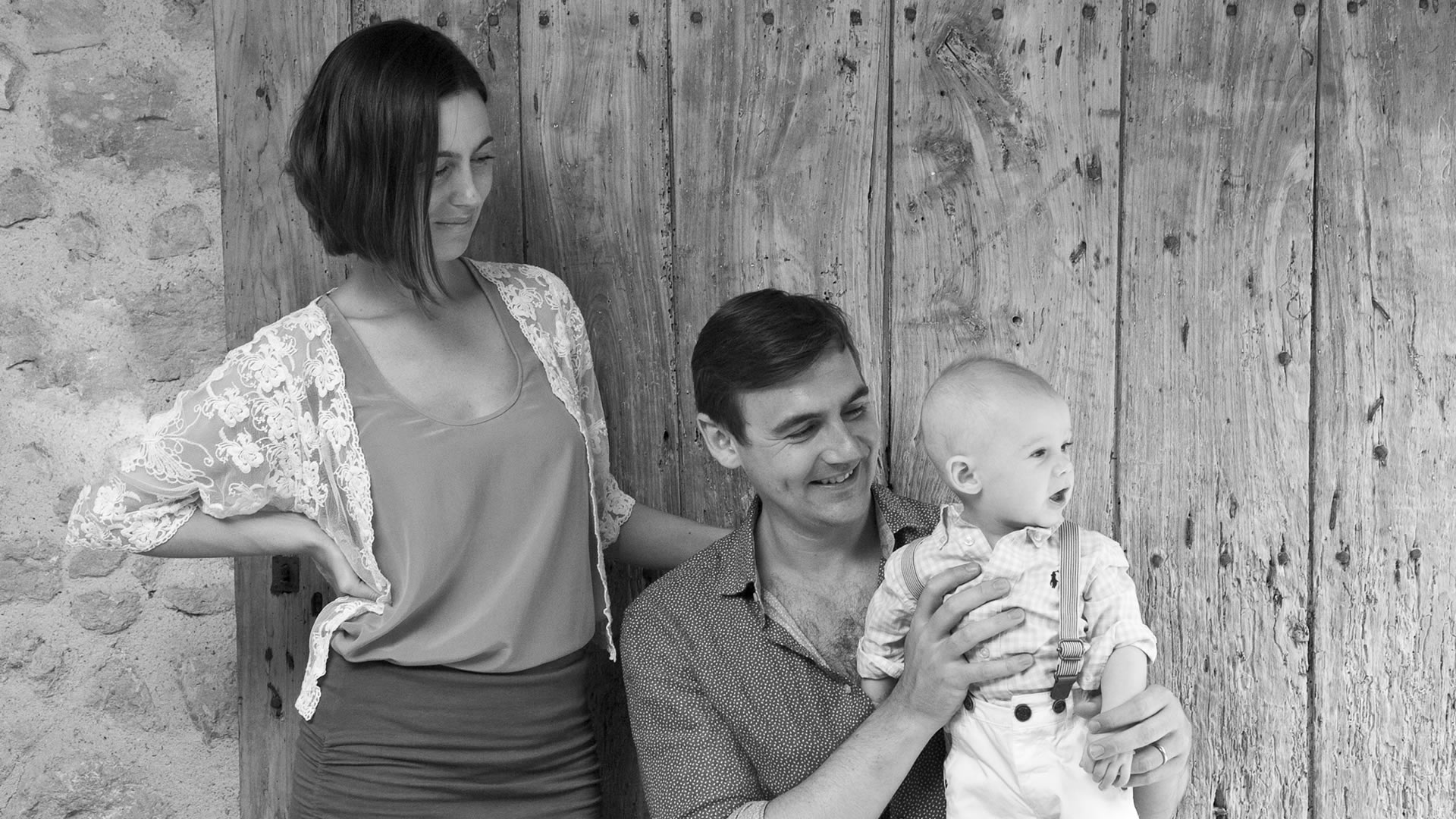 Portrait de famille en noir et blanc de parents avec leur bébé devant un mur en bois rustique – photographie de famille dans le Médoc, France