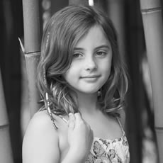 Portrait en noir et blanc d'une petite fille en robe fleurie, debout devant un fond de bambous.