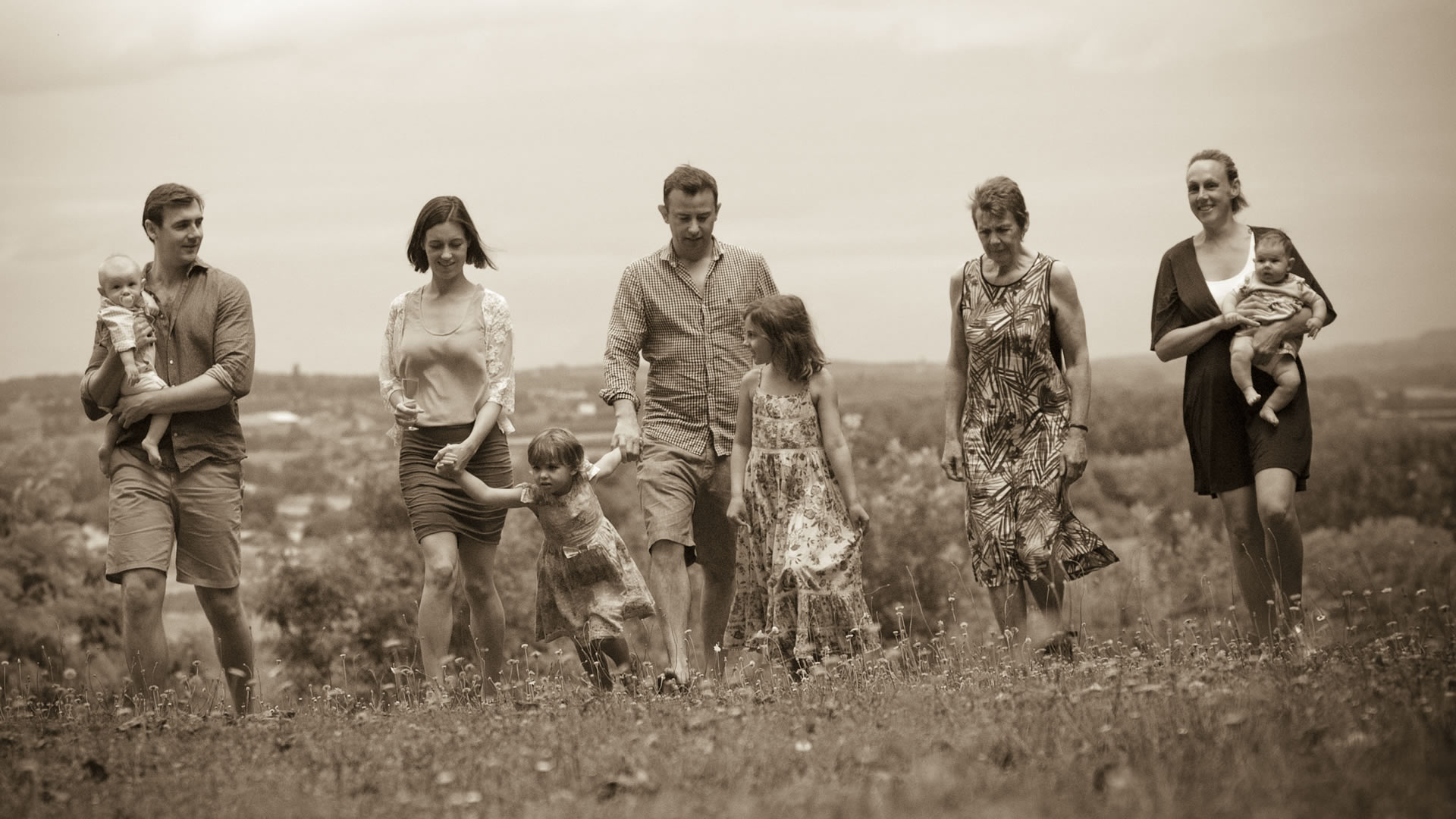 Portrait de famille aux tons sépia d'adultes et d'enfants marchant ensemble dans une prairie de fleurs sauvages.
