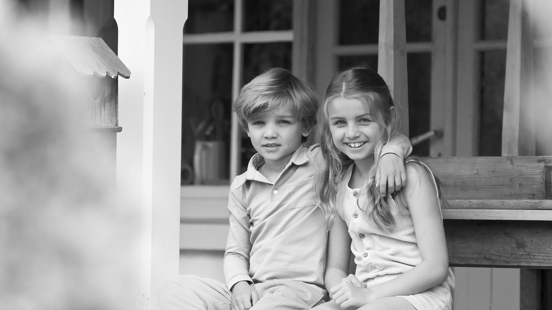 Portrait en noir et blanc d'un garçon et d'une fille souriants, assis côte à côte sur un banc de porche