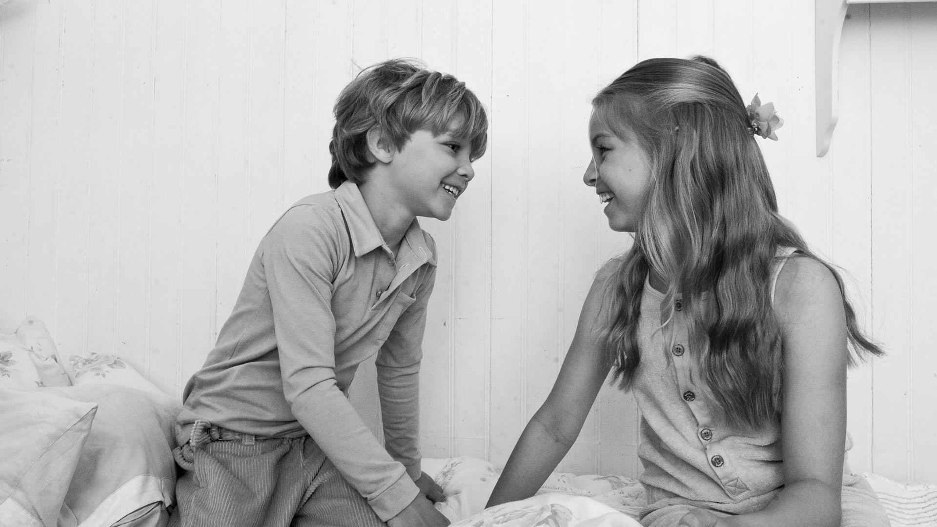 Photo en noir et blanc d'un garçon et d'une fille riant, face à face sur un lit devant un mur lambrissé blanc