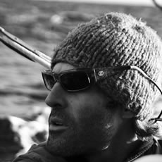 Gros plan en noir et blanc d'un homme barbu en bonnet en maille et lunettes de soleil, avec une eau agitée derrière lui